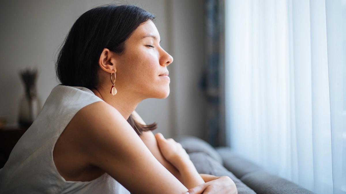 woman leans over couch with arms crossed and resting on the couch, eyes closed serenely, facing towards the light of a window