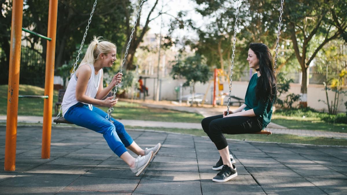 two women sitting next to each other on swings at park laughing