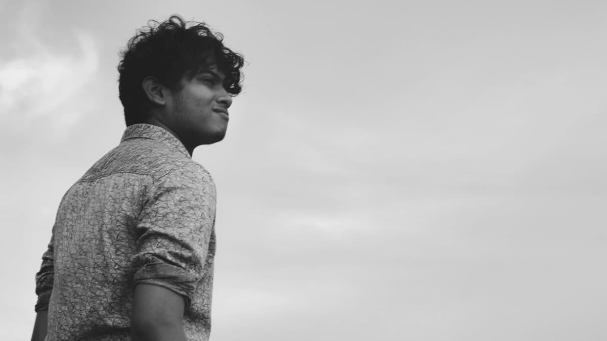 Young man with a button up shirt and rolled up sleeves looks off into the distance, clouds and sky are the background, black and white