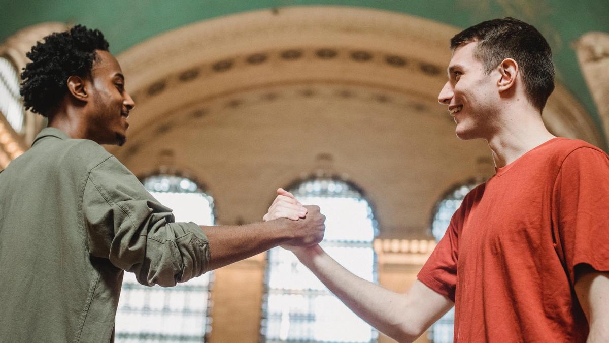 Two young men shake hands while smiling inside a large building with arch shaped windows. 