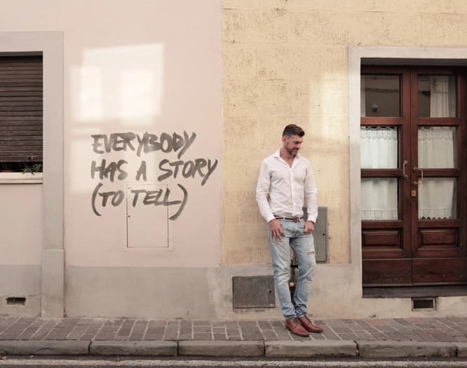 man standing against a wall with graffiti that says "Everybody has a story (to tell)"