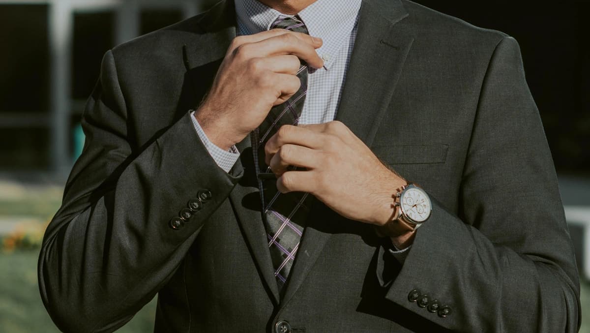 Man adjusts his tie; he has a black suit on and a watch, zoomed in on his torso and arms.